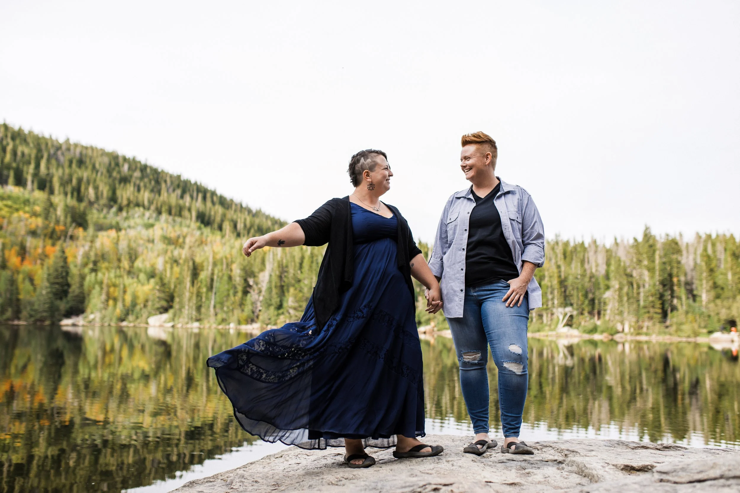 Two women holding hands and dancing on a rock by a lake, surrounded by trees and mountains, smiling at each other.