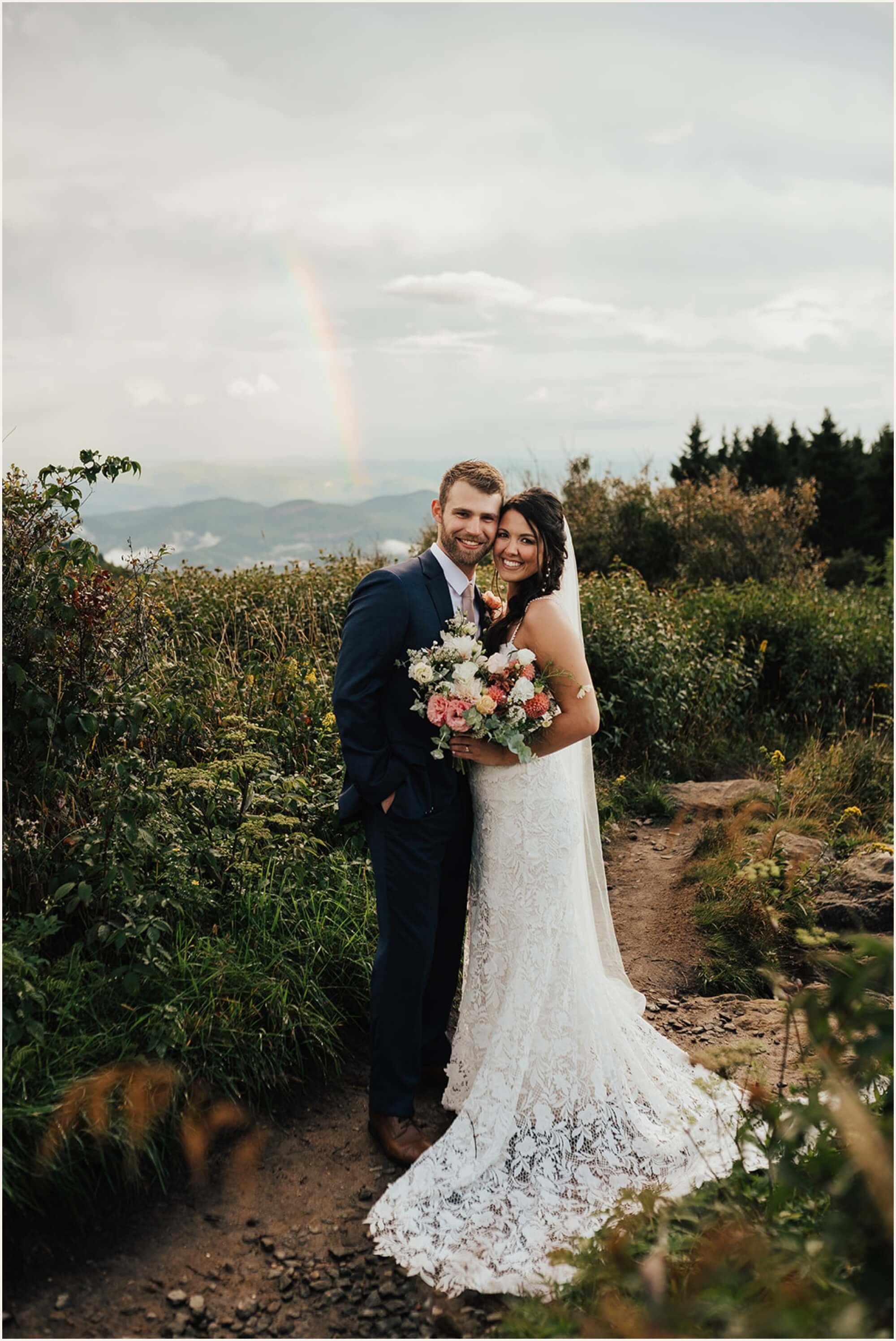 Bride and groom on mountain trail with rainbow behind them