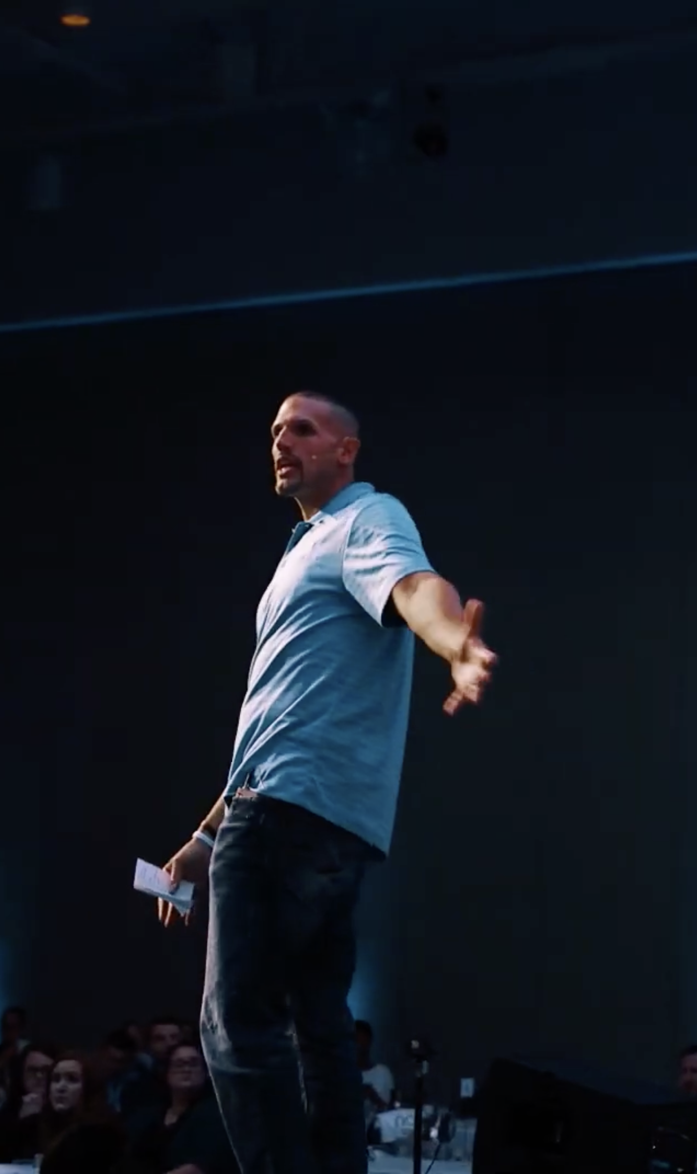 A man in a light blue shirt speaking passionately on stage with an audience watching.