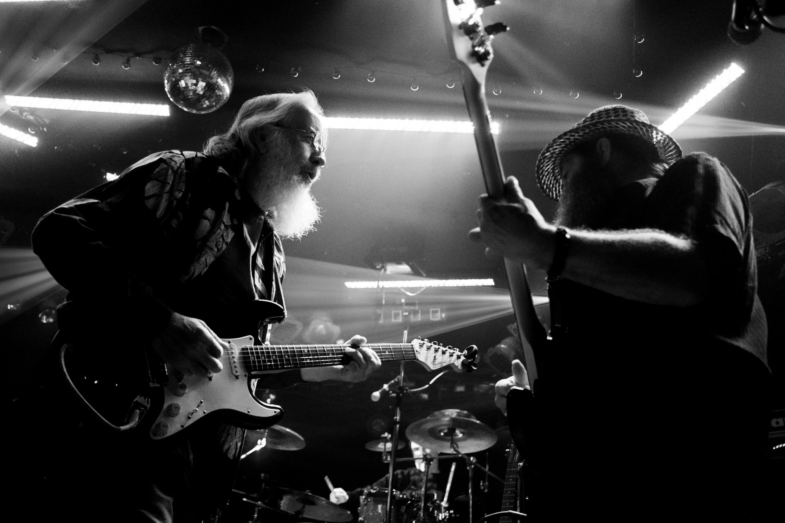 Two musicians perform on stage in a black-and-white photograph, one with long hair and a beard playing an electric guitar, and the other wearing a straw hat playing an electric bass guitar, with a drum set visible in the background.