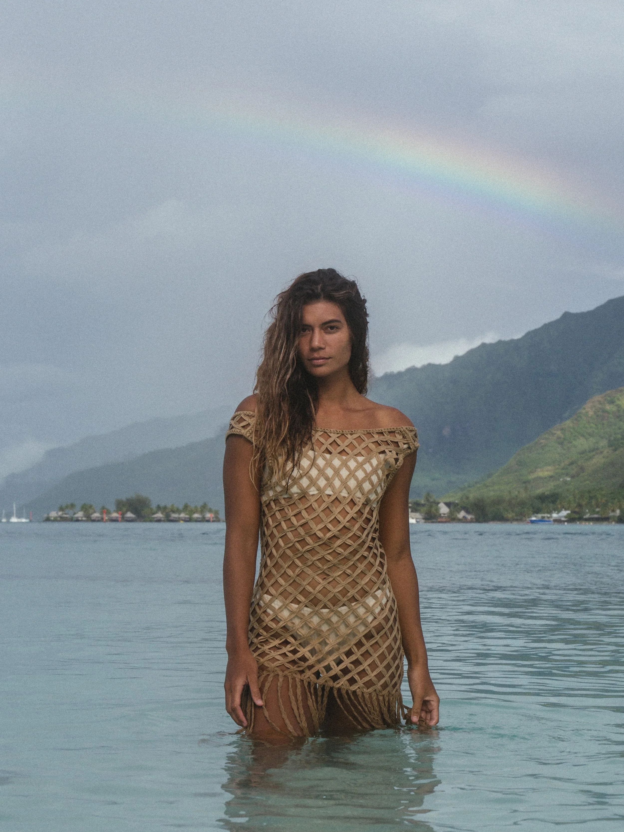 Model and Roxy Longboard Surfer, Vaimiti, wearing a brown cover up and bikini standing in a lagoon in Mo'orea with a rainbow and mountains behind her during a brand and fashion photoshoot with Warm Collective. 