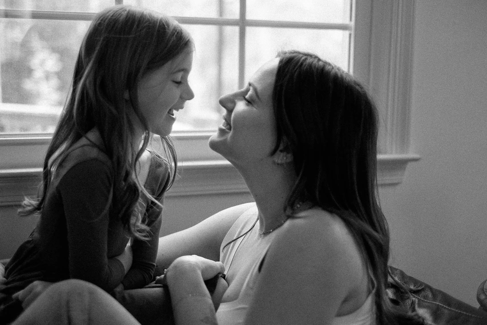 Mom and daughter smiling at each other while sitting on couch in front of window