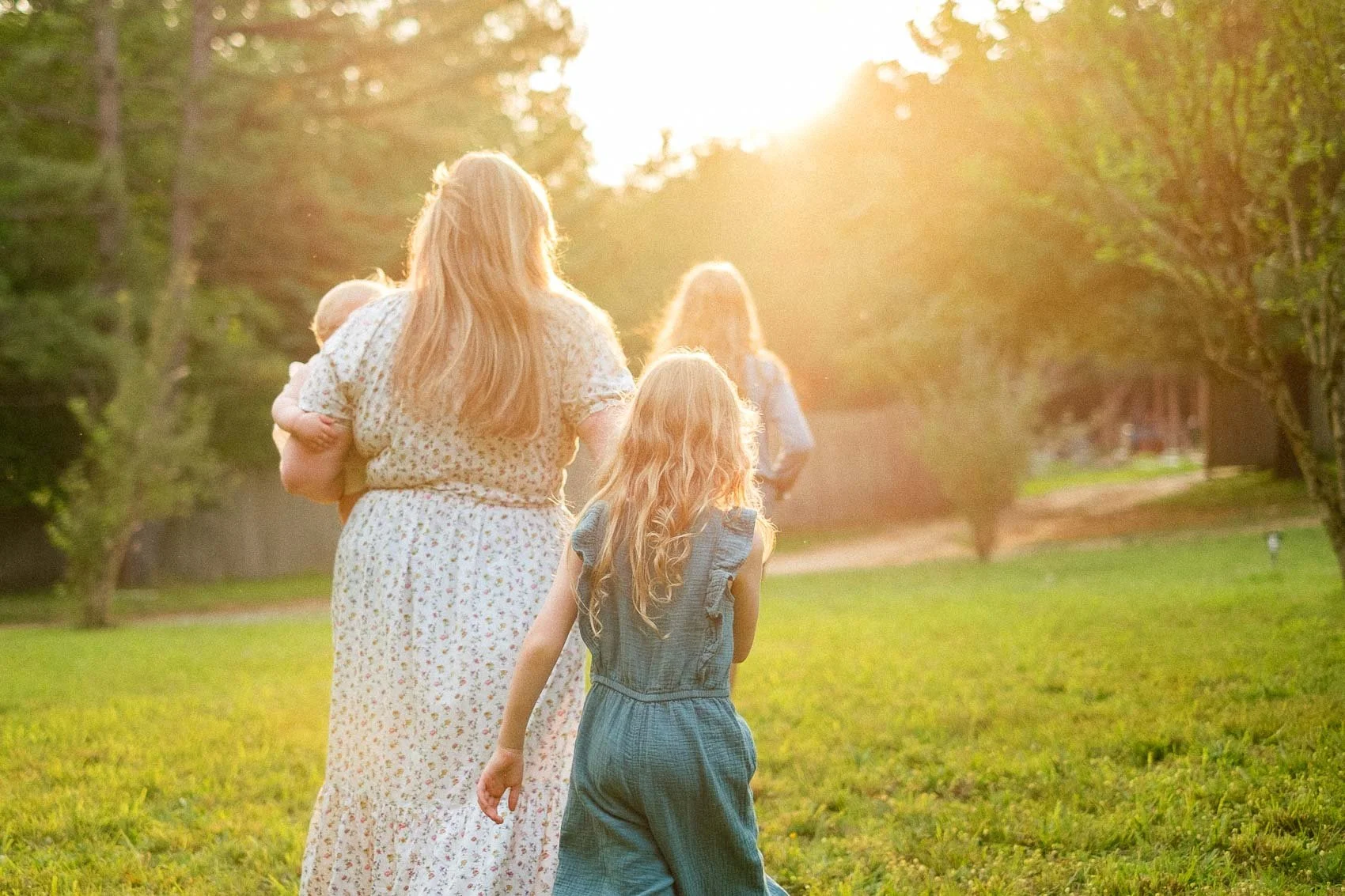 Mom holding baby and walking through green field towards glowing sunset with two older children