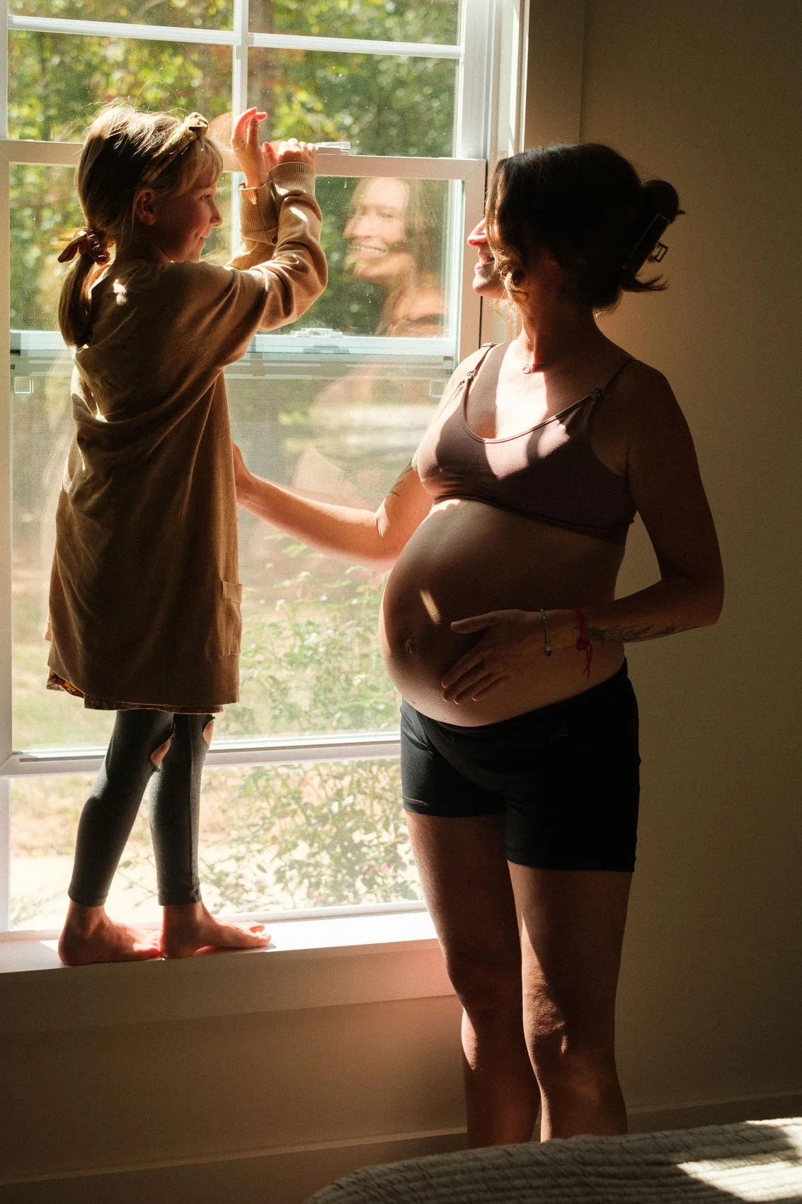 Pregnant mother and daughter smiling at each other in front of window