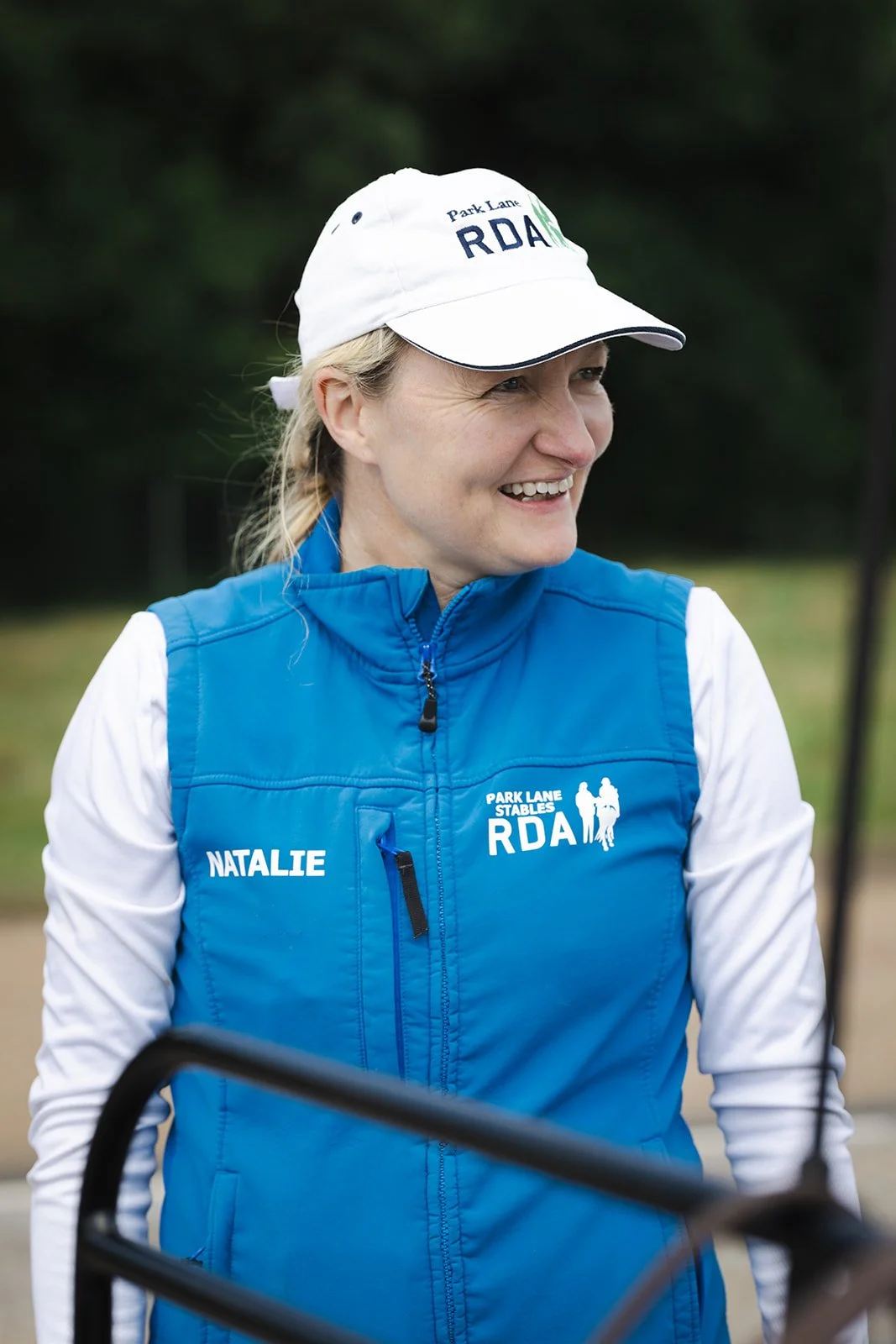 A woman with a braid, wearing a navy blue vest, gray and navy shirt, and a dark baseball cap with a logo, stands inside a wooden stable, smiling as she holds a light-colored horse's face close to hers.