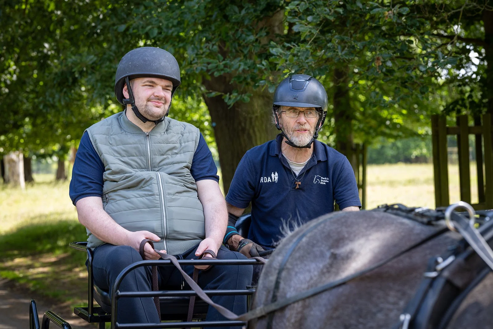 Two riding a carriage, both wearing helmets. One man with glasses and a goatee, and the other man with a beard. The background features green trees and grass.