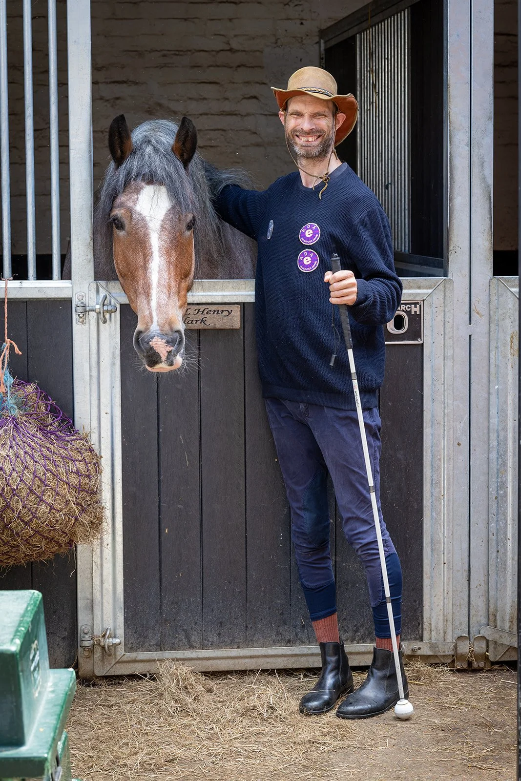 A man with a beard and blue eyes smiling, wearing a blue jacket and teal shirt, standing outdoors in front of a wooden and metal shed.
