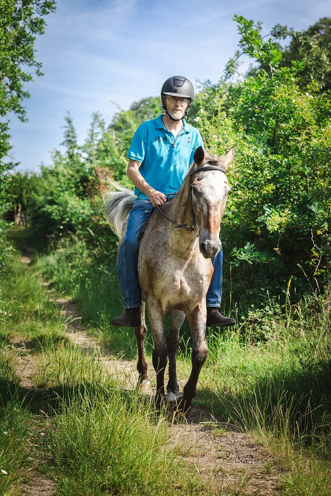 Smiling man with a beard and glasses wearing a flat cap, a gray blazer, and a scarf, sitting outdoors on a cloudy day with greenery and houses in the background.