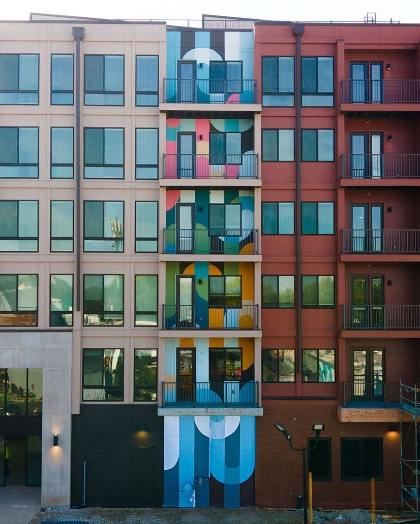 Multi-story building with colorful abstract mural on the central section, featuring circles and curved shapes in various colors, with balconies on each floor.
