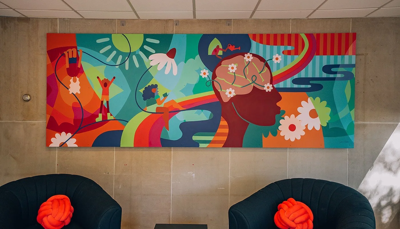 Colorful abstract mural with a silhouette of a human head, surrounded by images of people, flowers, rainbows, and abstract shapes, in a modern interior space.