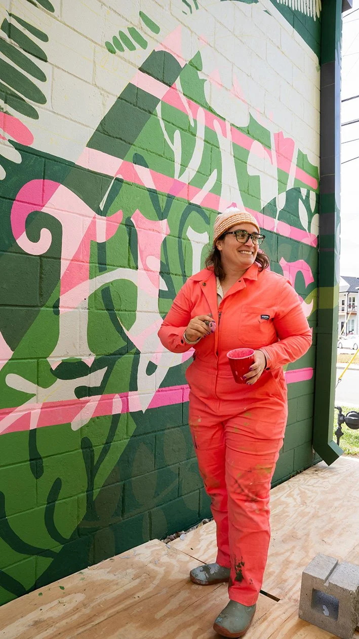 A woman wearing glasses, a beanie, and coral-colored work clothes stands in front of a colorful mural on a brick wall, holding a red cup and a paintbrush, smiling.