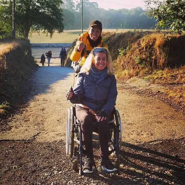 A woman in a wheelchair smiling and sitting on a dirt path outdoors, with a smiling man behind her giving a thumbs-up. They are dressed in outdoor clothing, and other people are walking in the background on a path surrounded by trees and grassy hills.
