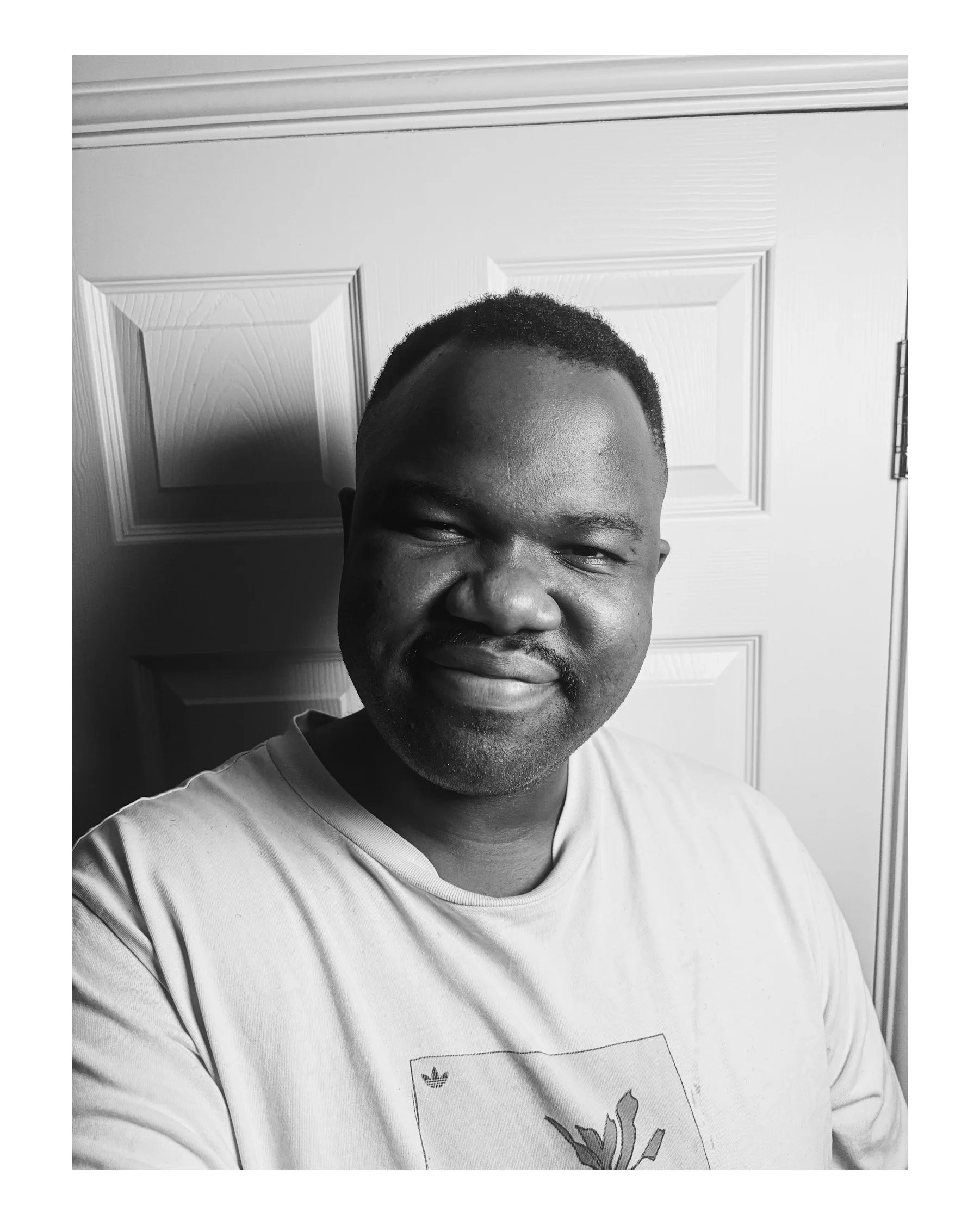 Black and white photo of a smiling man wearing a shirt with a graphic on it, standing in front of a paneled door.
