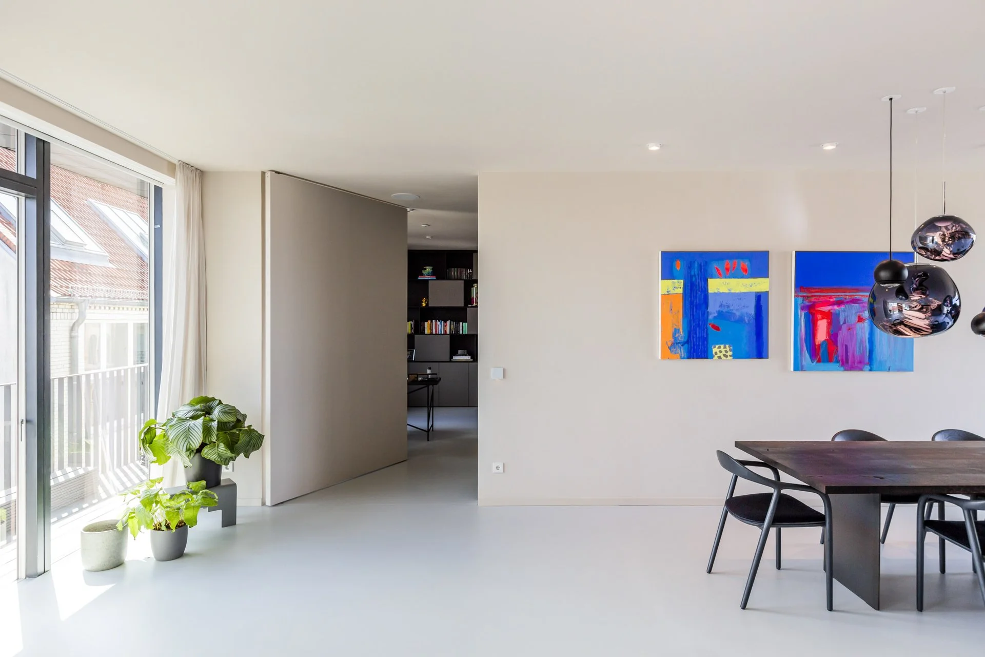 Moderner, heller Wohnraum mit bunten abstrakten Wandbildern, schwarzem Esstisch mit vier schwarzen Stühlen, mehreren Pflanzen neben einer großen Glas-Tür zur Terrasse, und schwarzer Bücherregal im Hintergrund.