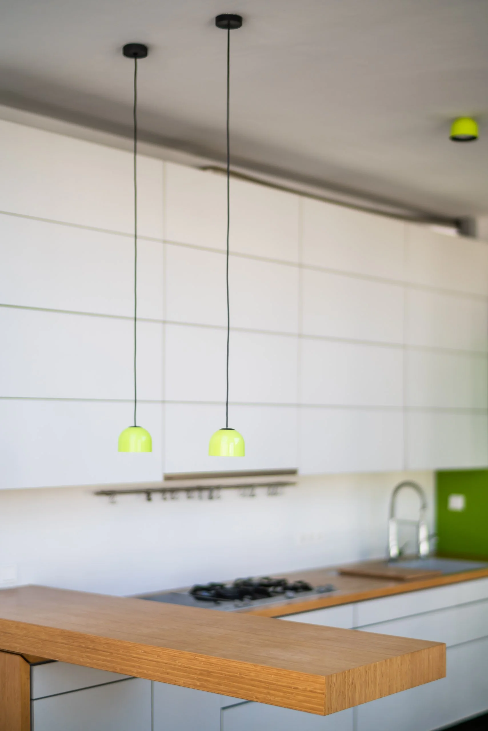 Moderne Küche mit weißen Schränken, Holzarbeitsplatte, grüner Rückwand, Zwei Hängelampen mit grünen Lampenschirmen, Spüle und Gasherd.