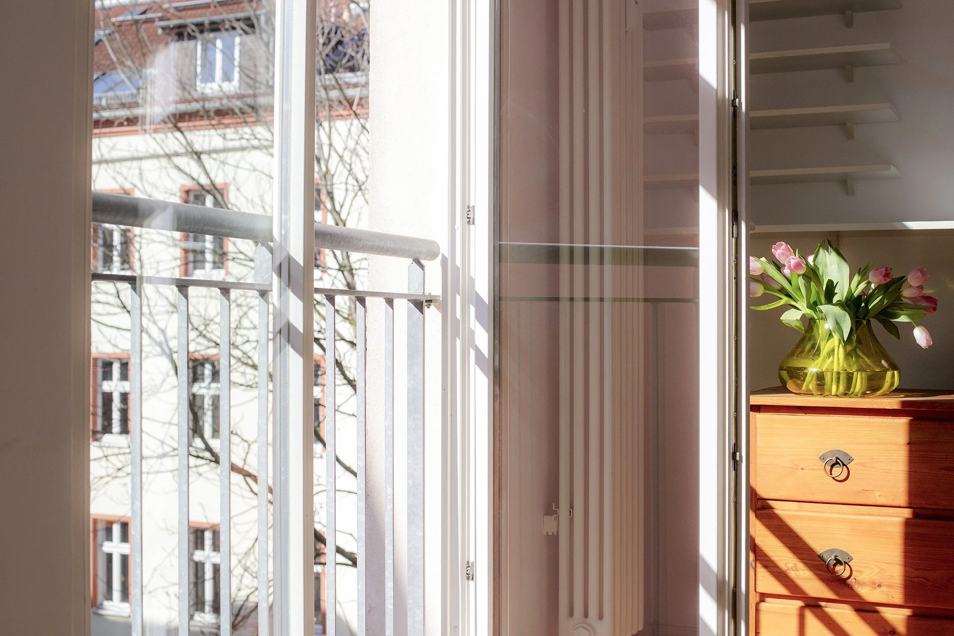 Blick aus einem Fenster auf einen Balkon mit Zinnen, im Inneren ein Holzkommode mit einem Blumenvasen voller Tulpen, Sonnenlicht fällt ins Zimmer.
