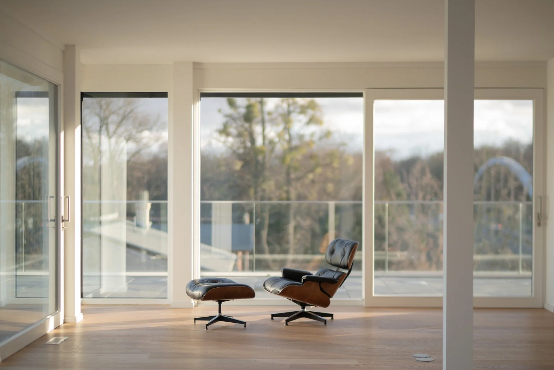 Moderner Raum mit großen Fenstern und einem stilvollen braunen Ledersessel mit Hocker, Blick nach draußen auf eine ruhige Landschaft mit Bäumen, Holzboden.