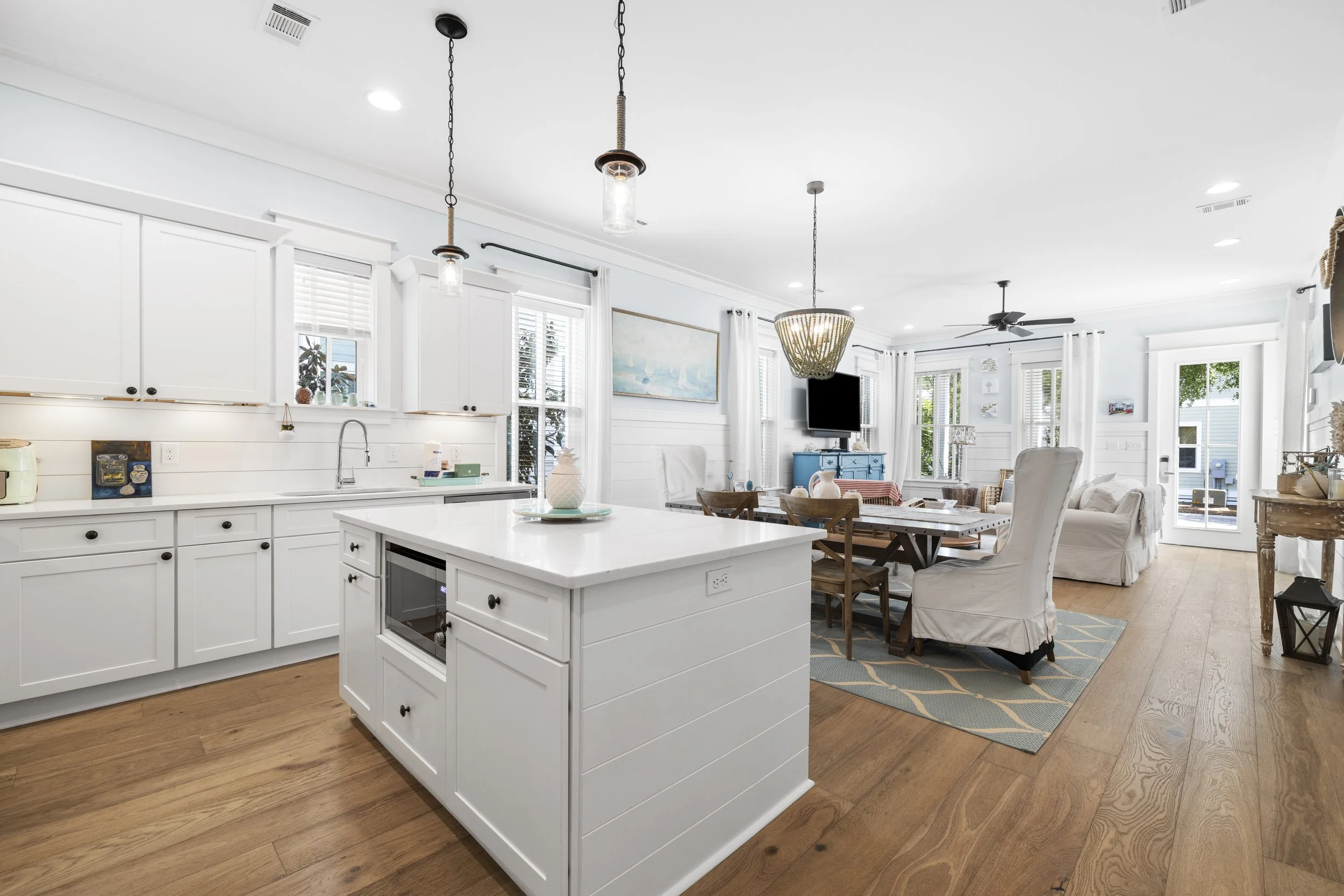 Open-concept kitchen and living room with white cabinetry, island, dining area, and seating, featuring natural light, wood floors, and coastal decor.