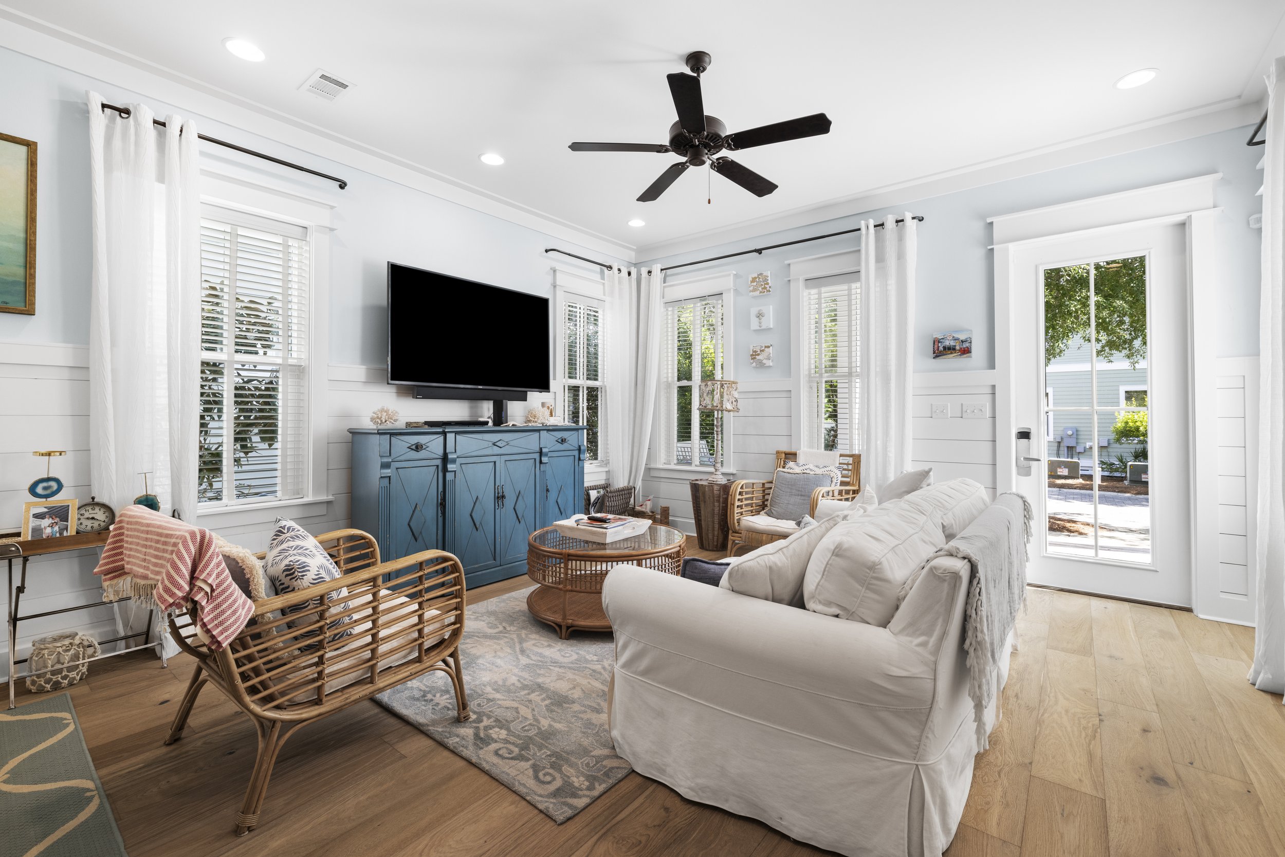 Light-filled living room with white walls, white curtains, light wood flooring, a white sofa, rattan chairs, a blue cabinet, a flat-screen TV, and a glass door leading outside.