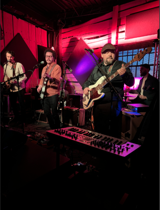 A band performing live on stage with four members playing guitars and singing, in a cozy, warmly lit wooden venue.
