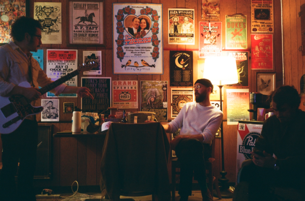 A man playing an electric guitar to a small audience in a cozy, wood-paneled room decorated with vintage concert posters.