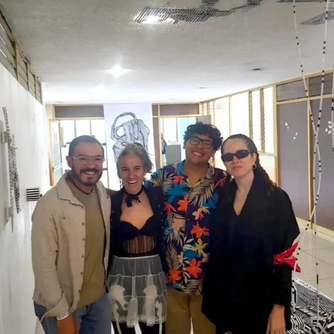 Four friends smiling and standing together indoors, with artwork on the wall behind them and decorative panels to the side.