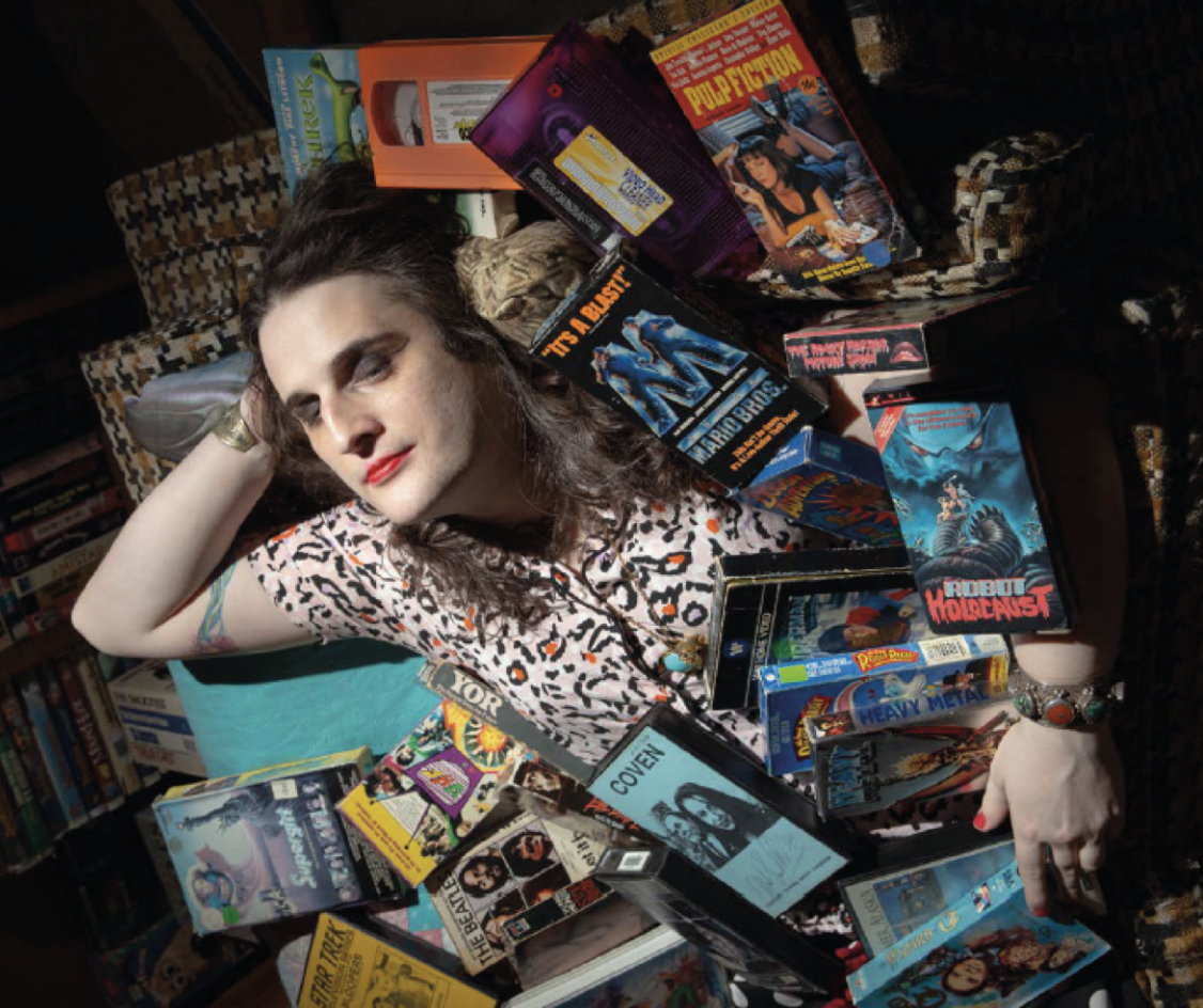 Person lying on a pile of vintage VHS tapes and books in a bookstore or library.