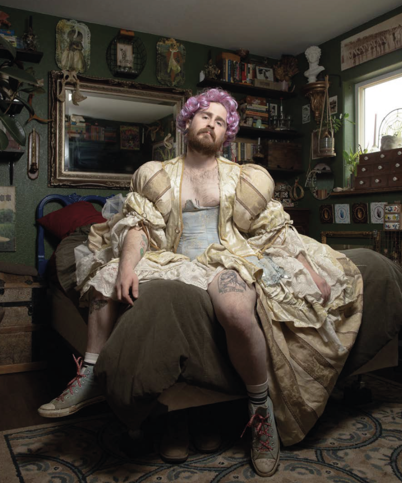 A person dressed in an ornate, historic-style yellow dress with puffy sleeves, sitting on a bed in a cozy, decorated bedroom. They are wearing purple curly wig, sneakers, and striped socks, with tattoos visible on their legs. The room has green walls, a large mirror, bookshelves, plants, and various decorations.