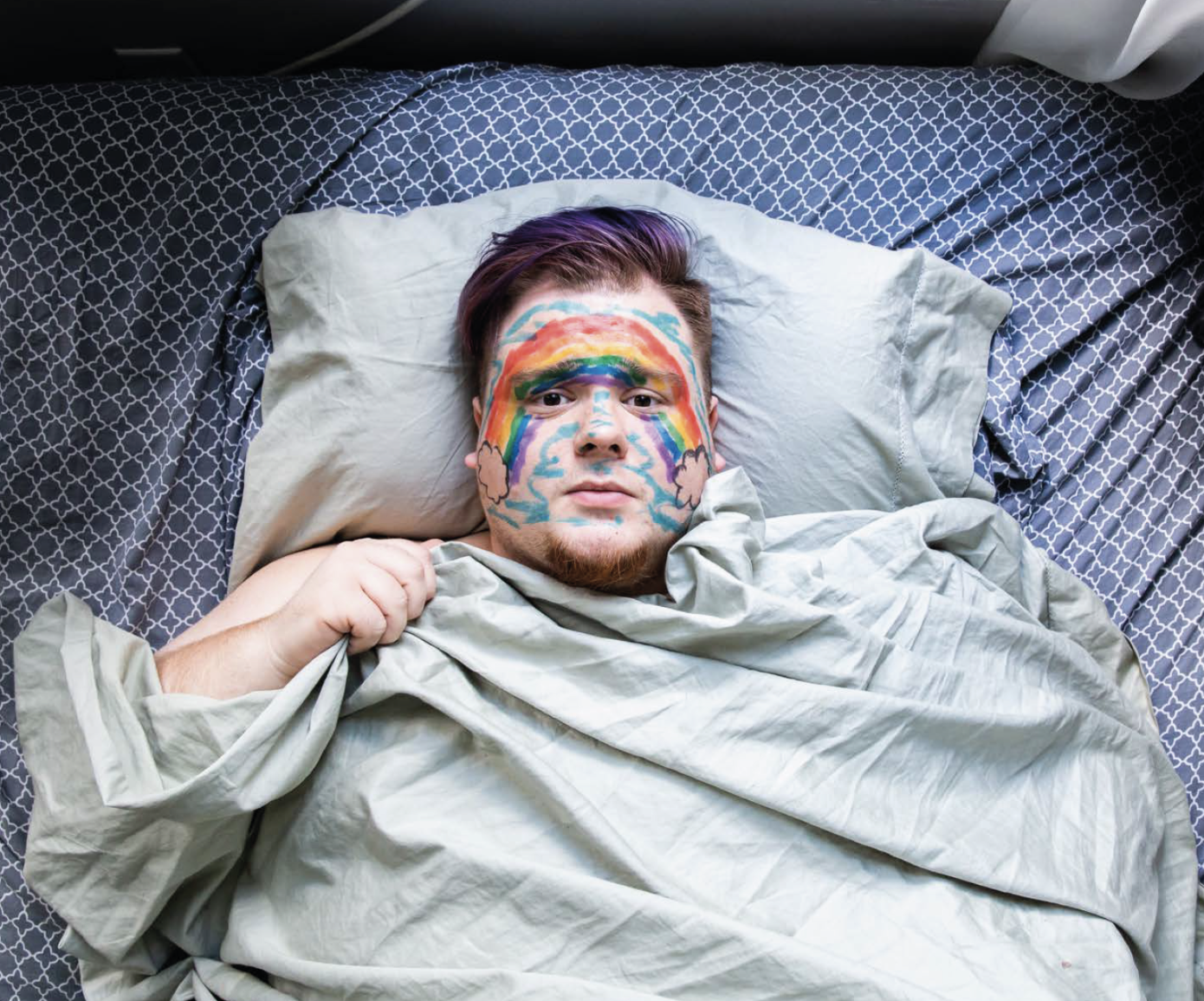 A man lying in bed with rainbow face paint, showing a rainbow and cloud design on his face.