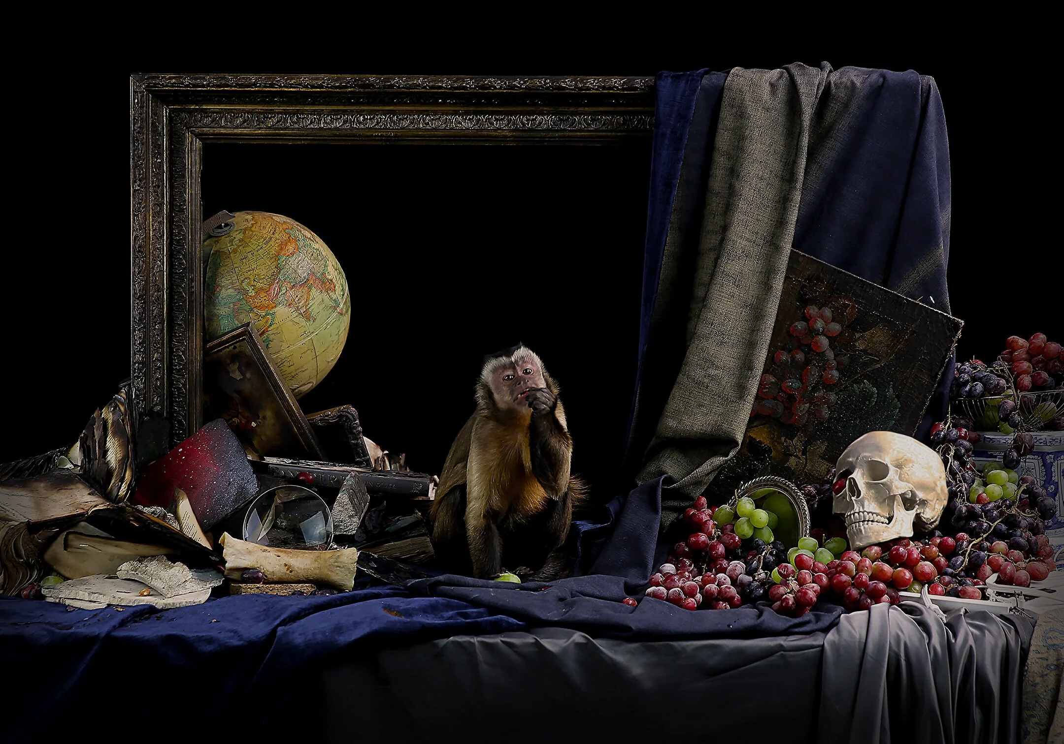 A cluttered scene with an open ornate frame, a globe, a monkey holding something to its mouth, scattered books, a skull, grapes, and dark drapery against a black background.