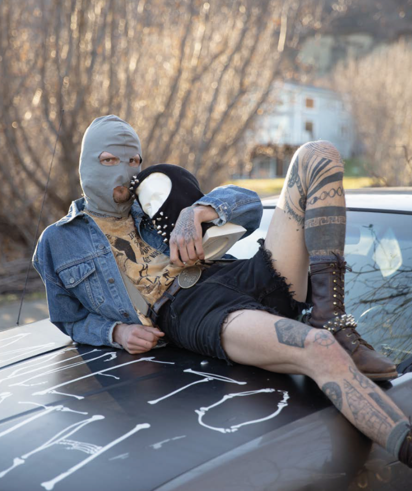 Person wearing a gray ski mask, denim jacket, and black shorts, with tattoos, is lying on the hood of a car, holding a smaller person wearing a hood and mask. The scene is outdoors with a blurred background of trees and a house.