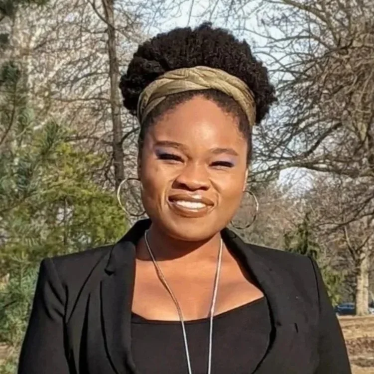 BIPOC therapist in DC standing outdoors, smiling, wearing a black blazer and necklace. Trees and greenery in the background.