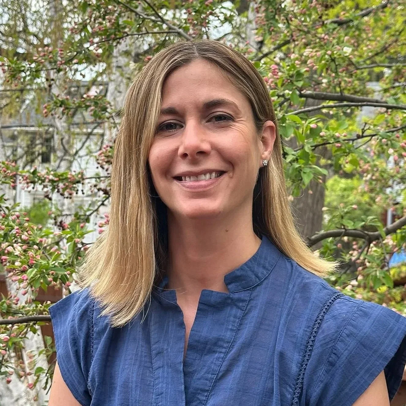 Experienced Trauma therapist in DC smiling in a blue top, standing outdoors with a backdrop of flowering branches and trees.