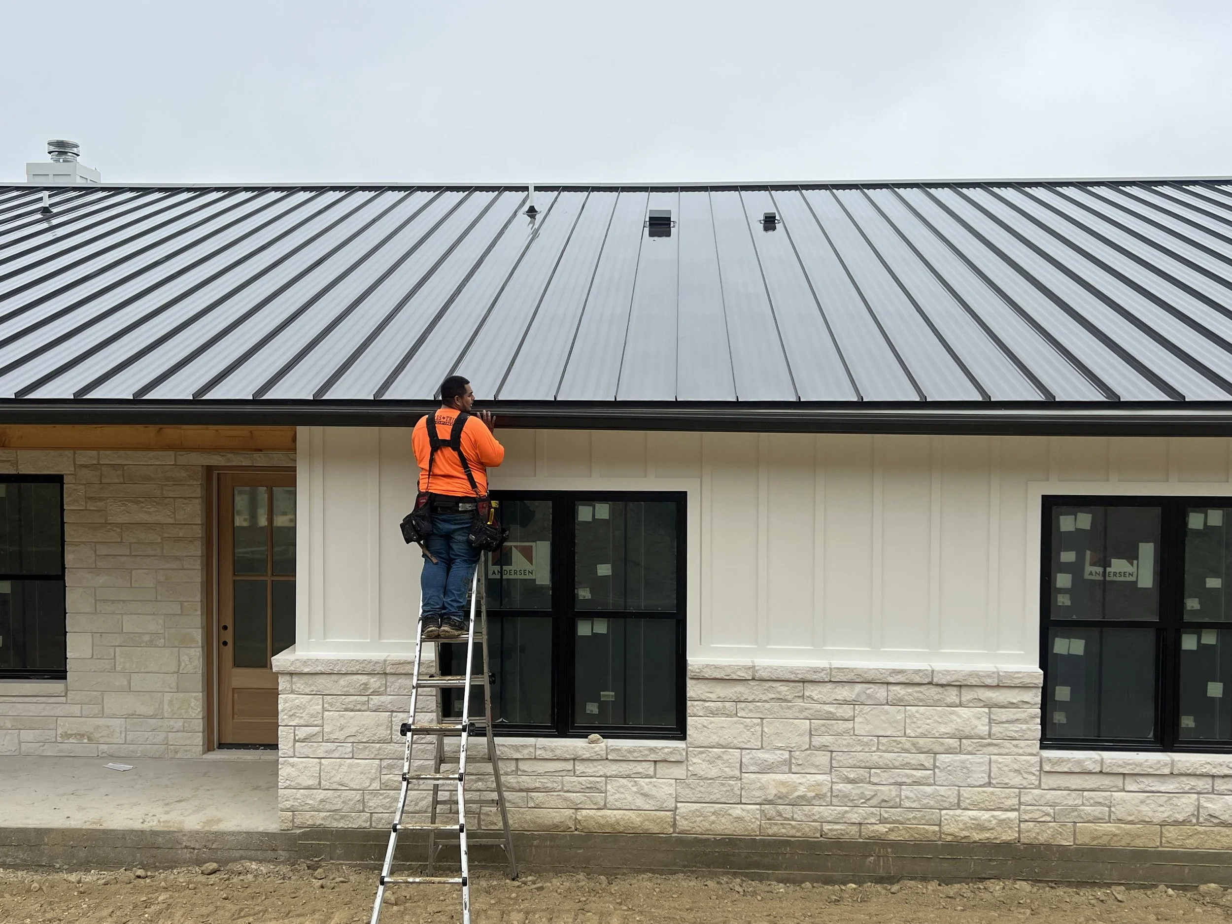A worker in an orange shirt standing on a ladder installing or inspecting a metal roof on a building with white siding and stone accents.