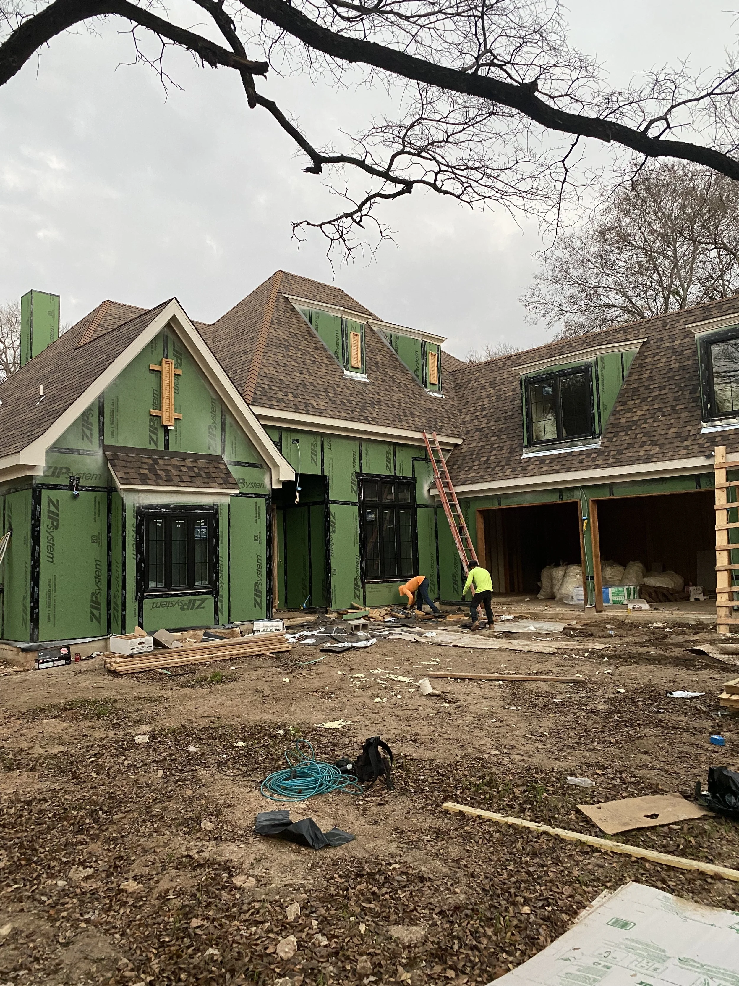 Construction workers working on the exterior of a house that is under construction, with green sheathing and brown roofing, in a yard with construction materials and equipment.