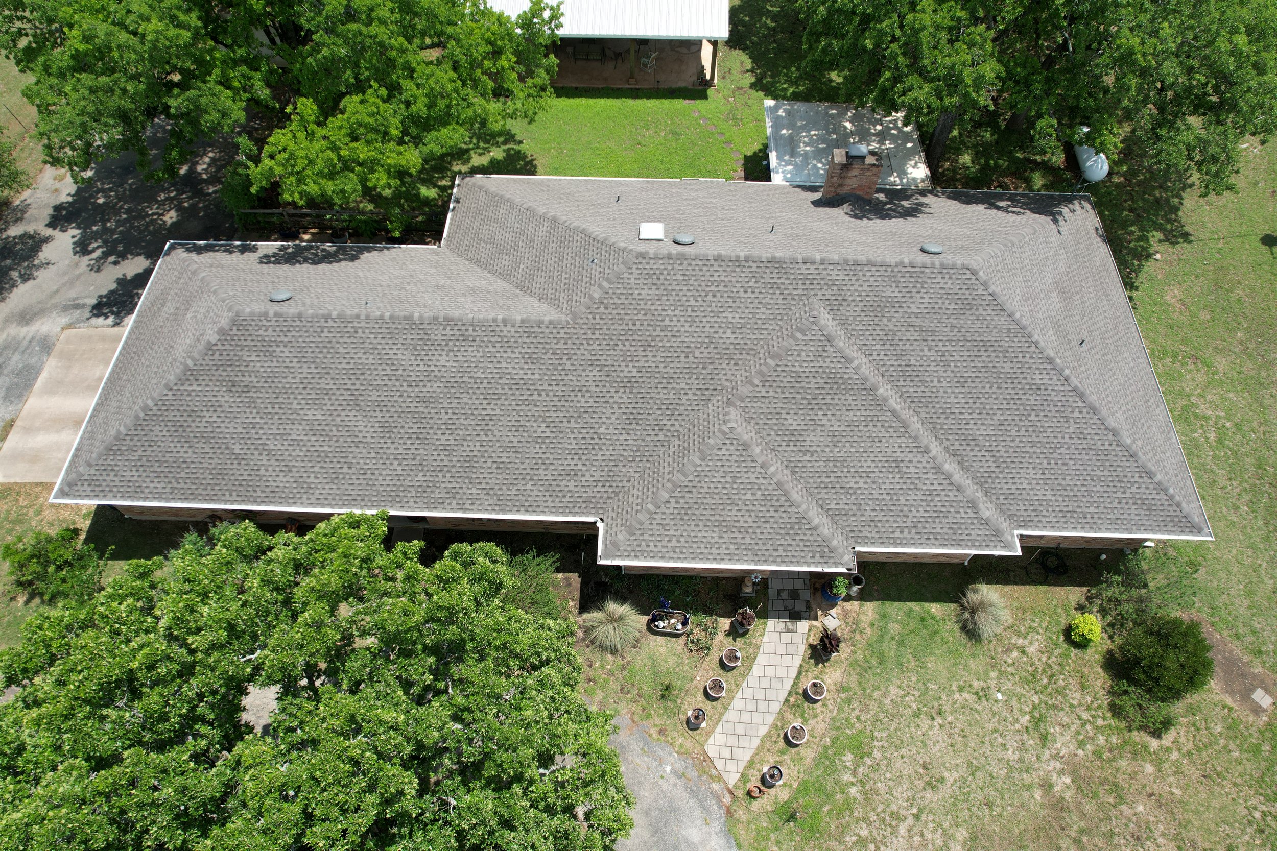 Aerial view of a house with a grey shingle roof, surrounded by green trees and a grassy yard. There is a small paved walkway leading to the front entrance, with potted plants along its sides. A driveway is visible on the left side of the house.