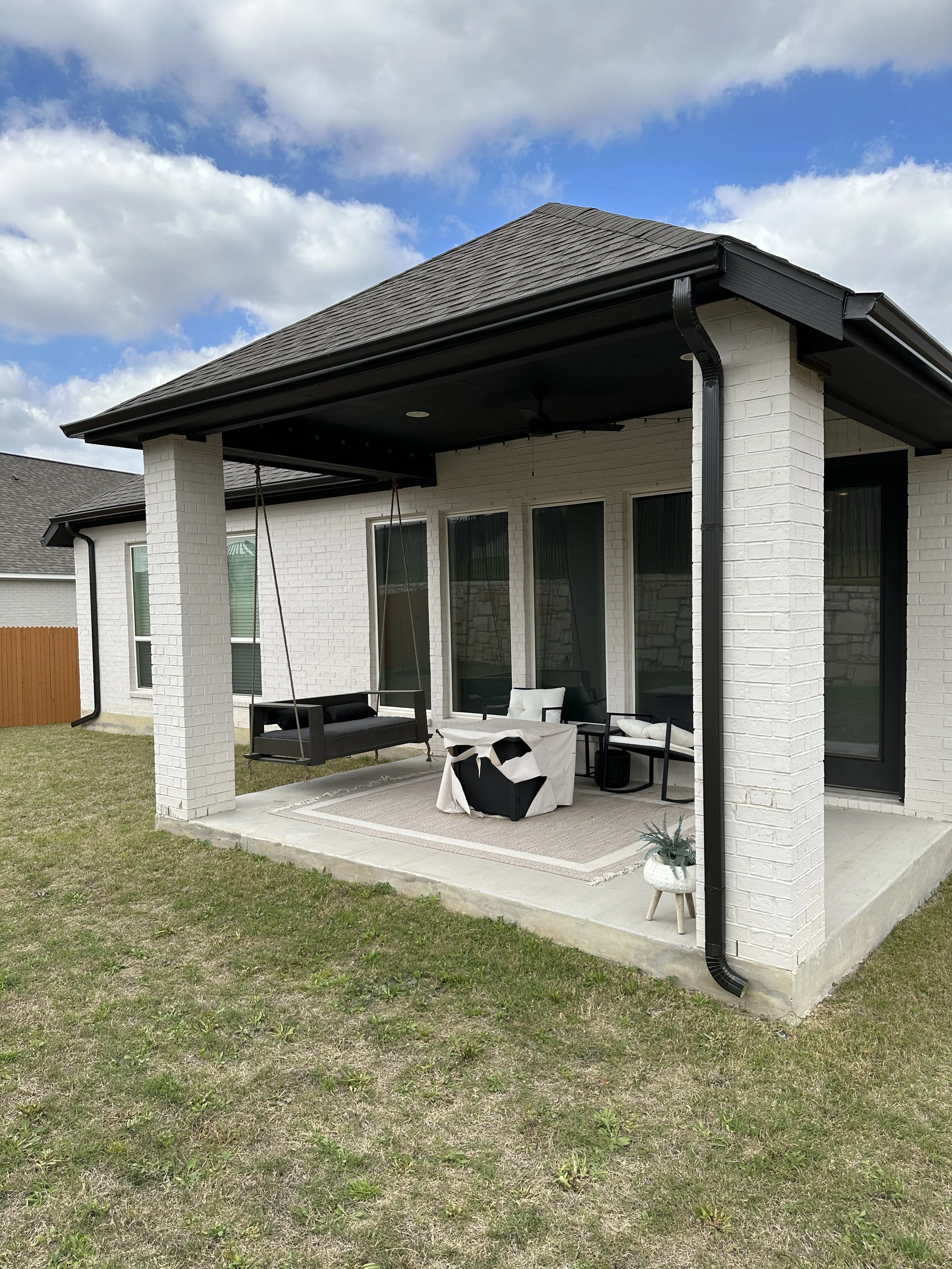 Back patio with hanging porch swing, outdoor furniture including chairs and a covered table, rug, and potted plant, attached to a white brick house with black trim, gray roof, and blue sky with clouds.