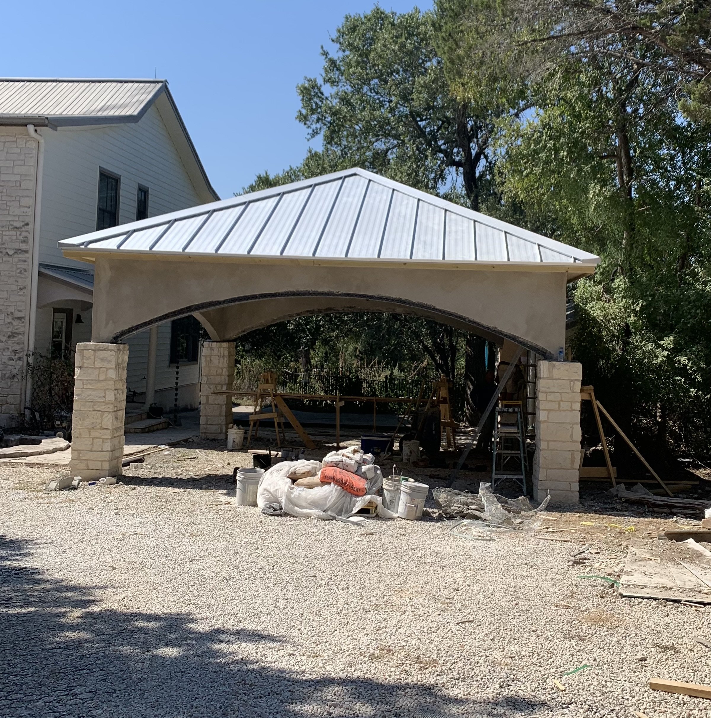 Under construction pavilion with metal roof, brick columns, and scaffolding, in a yard with gravel ground, construction tools, and supplies.