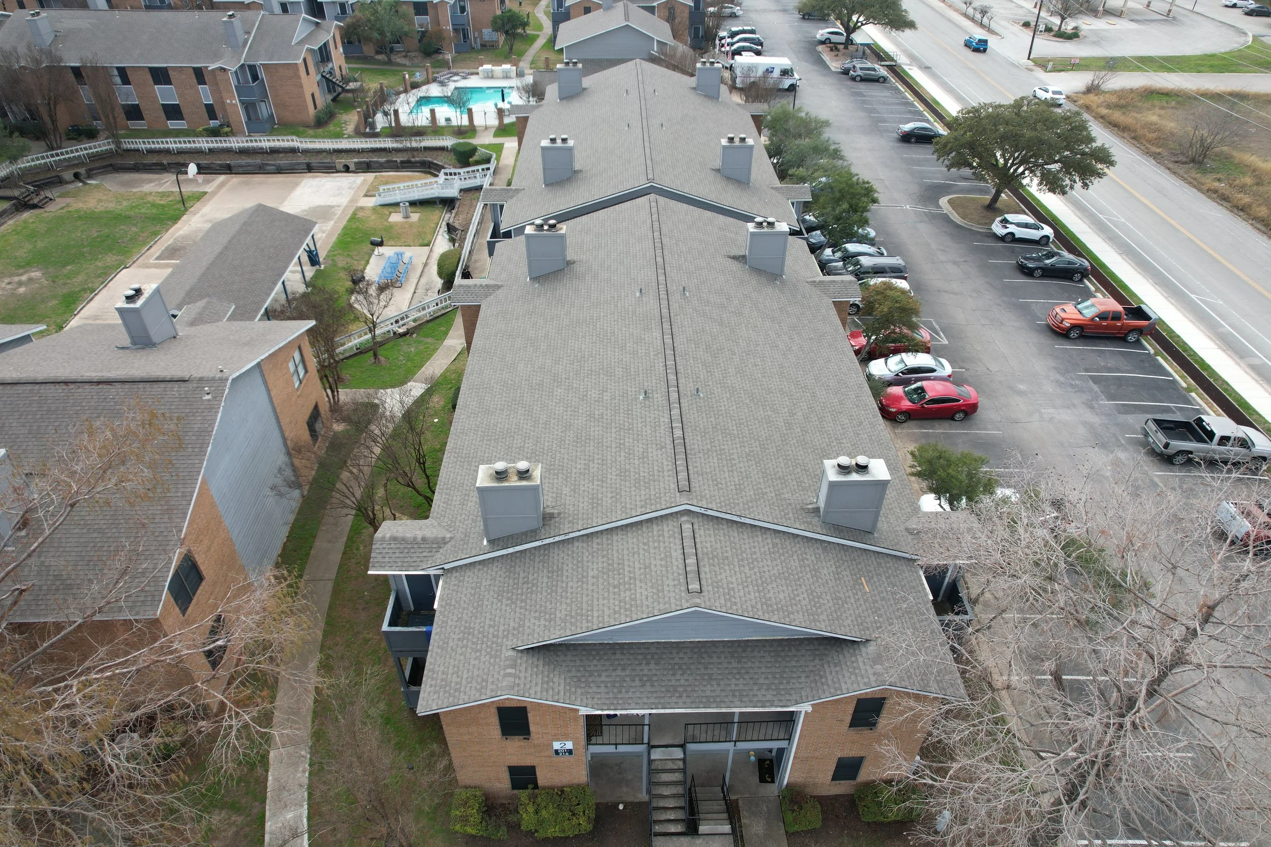 An overhead aerial view of a residential apartment complex with multiple buildings, parking lot, a swimming pool, tennis court, and walking paths.