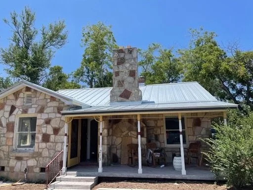 A stone house with a metal roof and a chimney, surrounded by trees, with a porch featuring chairs and a small table.
