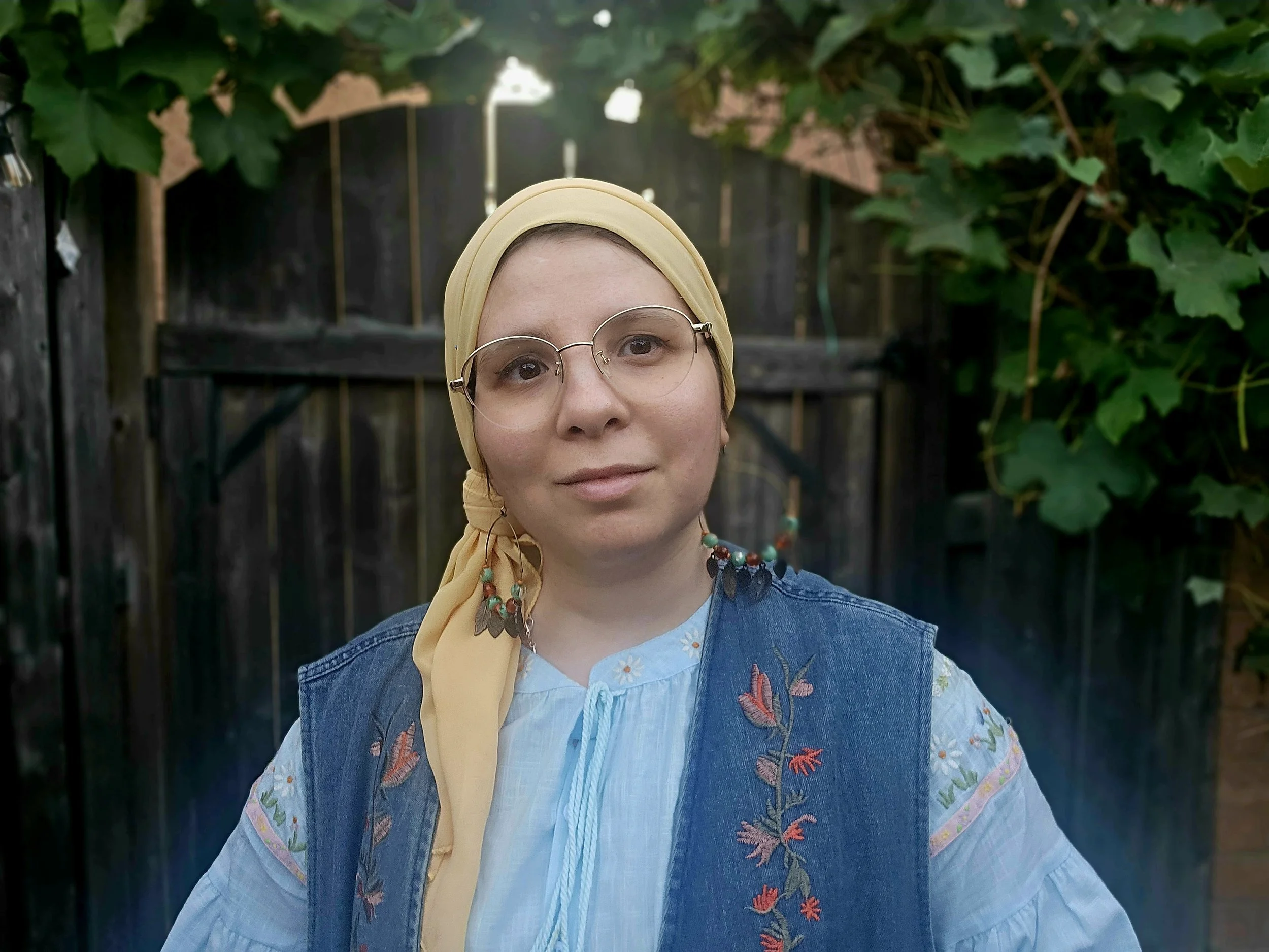 A woman with fair skin, wearing a yellow hijab, round glasses and oriental earrings, is standing in front of a wooden gate with leafy green plants overhead. She is dressed in a light blue embroidered blouse and a denim vest with floral embroidery.