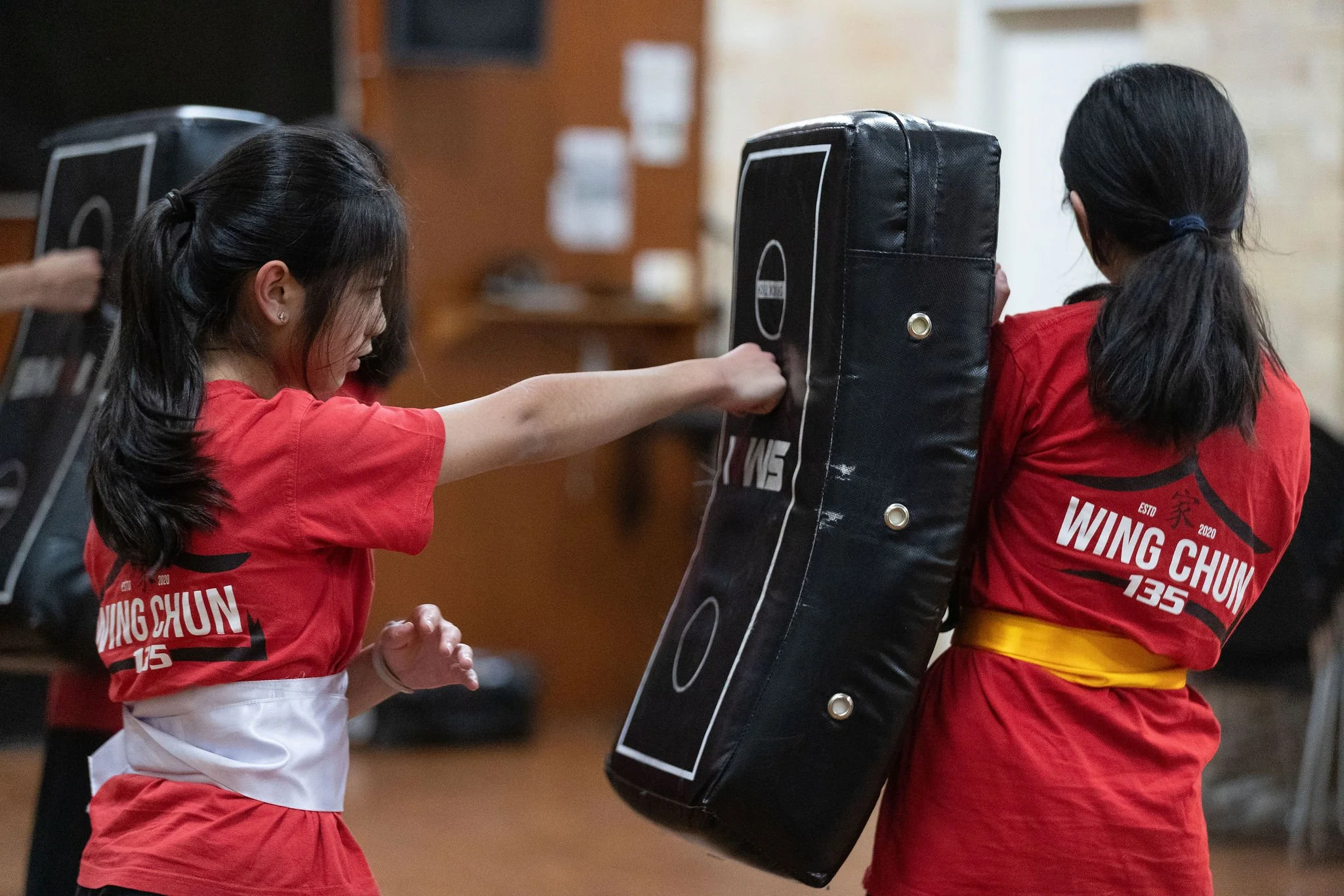 Two young women practicing Wing Chun kung fu martial arts in a training room, one is punching the padded shield held by the other.