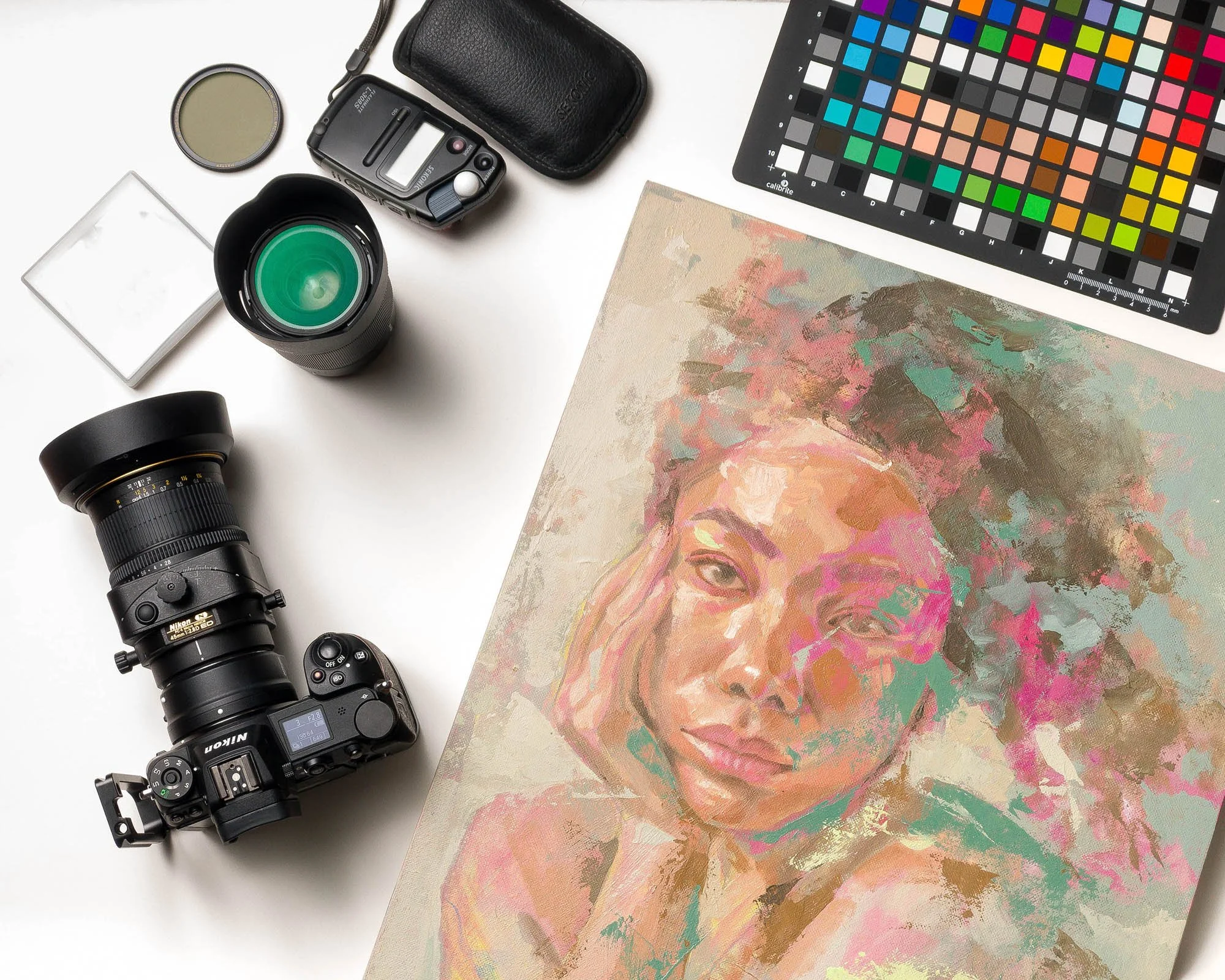 Photography equipment including camera lenses, a camera, a lens cap, a color calibration chart, a color palette, a small square container, a black case, and an abstract painted canvas, arranged on a white surface.