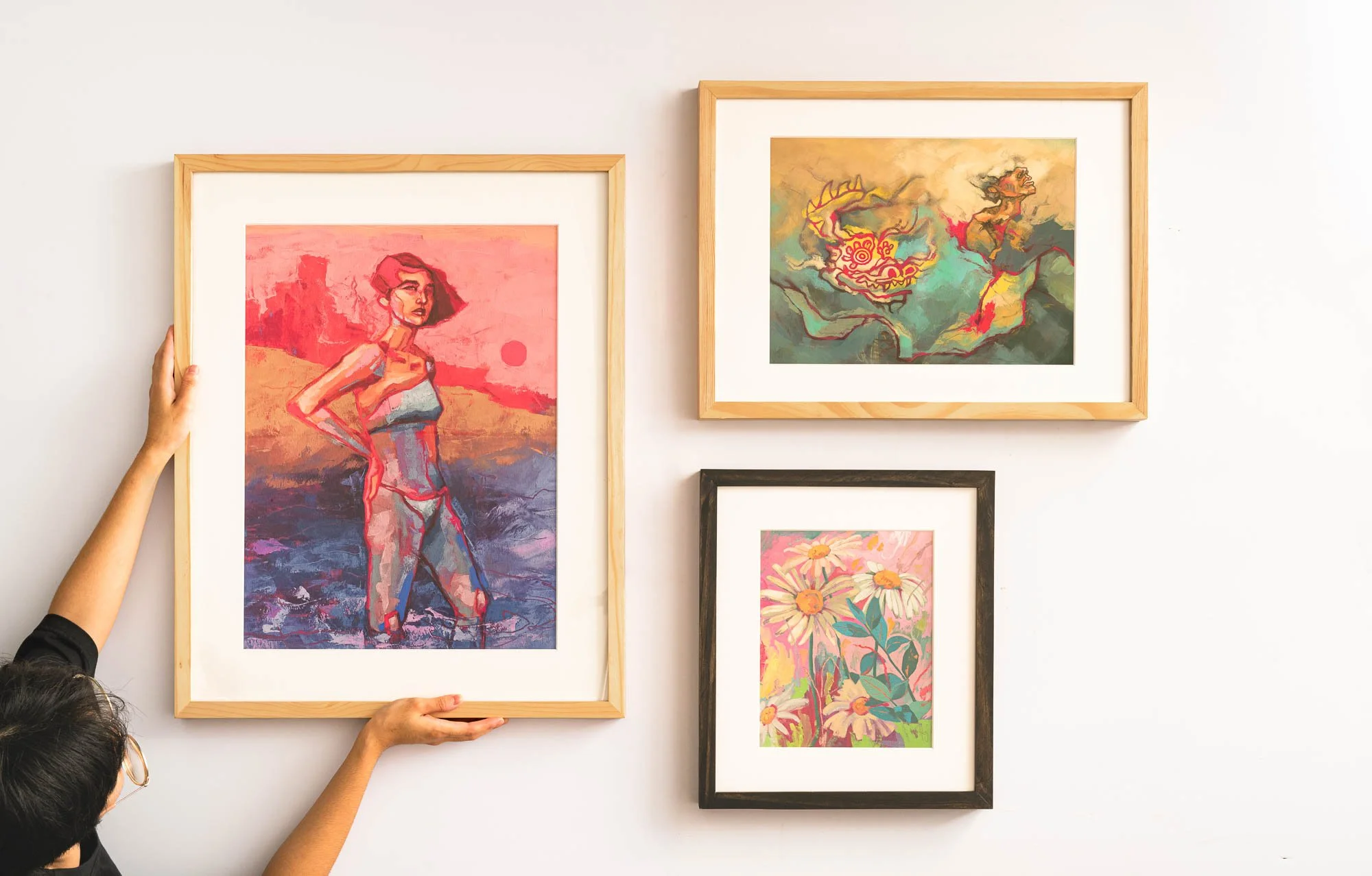 People hanging framed colorful artwork on a white wall, including a woman holding a painting of a woman in a landscape, and abstract and floral paintings.