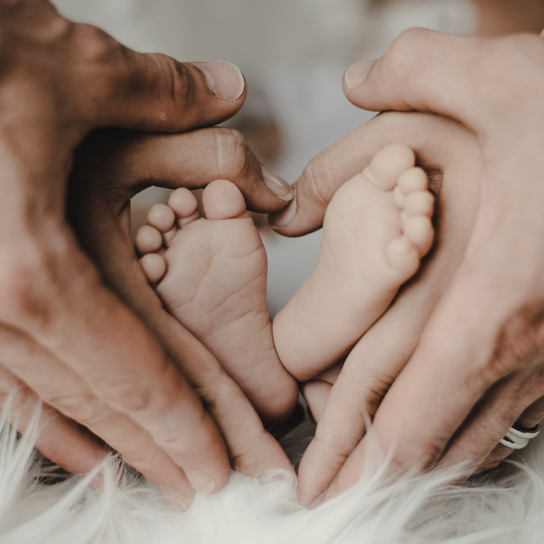 Close-up of adult hands holding a baby's feet, forming a heart shape, on a soft white surface.