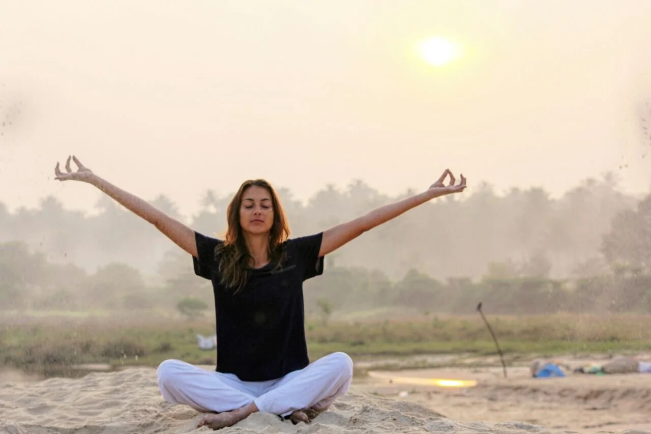 Femme méditant en position de lotus sur le sable, les bras ouverts, avec un paysage naturel et un coucher de soleil dans le fond.
