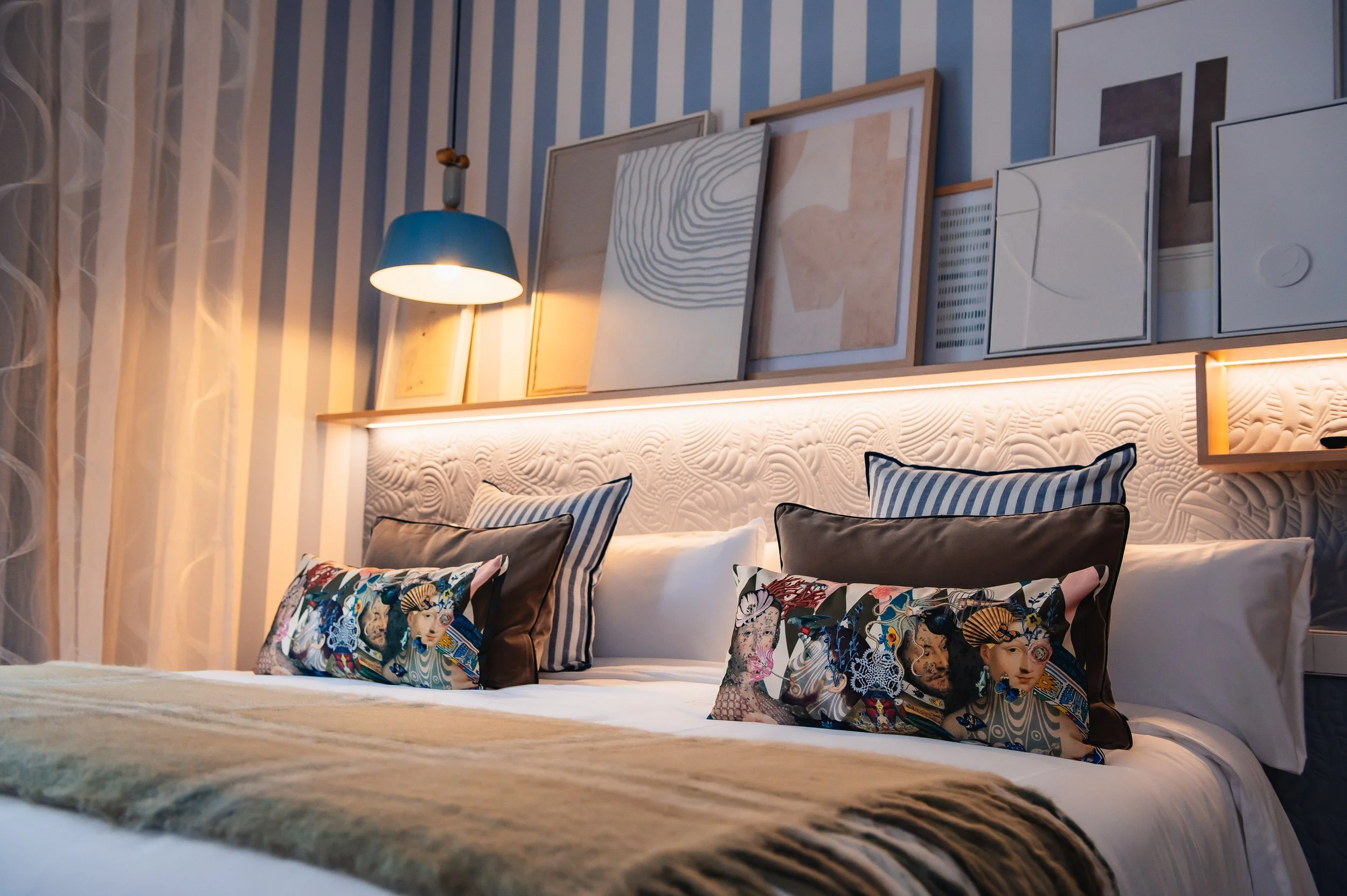A bed with decorative pillows in a room featuring striped walls, paintings, and a reading lamp
