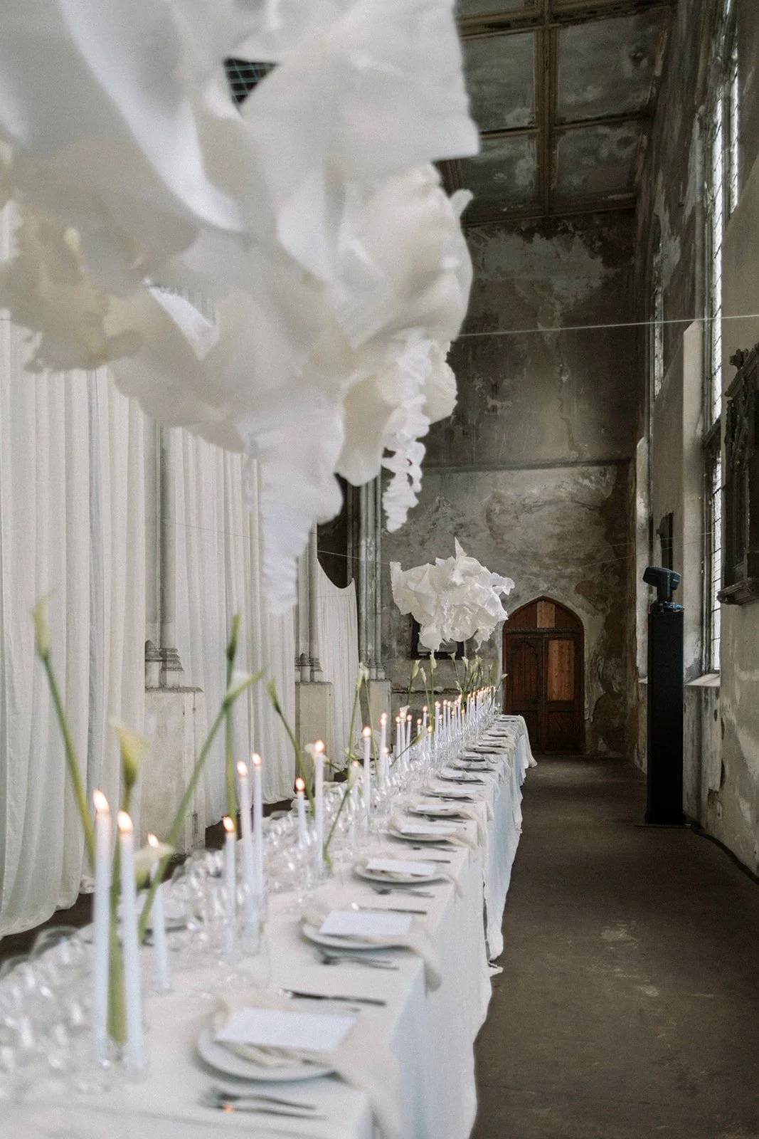 Long banquet table at Heritage Arts Centre, Bow, London, with white callas in glass vases, candles, in an rustic, gothic building in East London. Long crepe paper installations above the tables for a forward thinking, luxury and creative couple