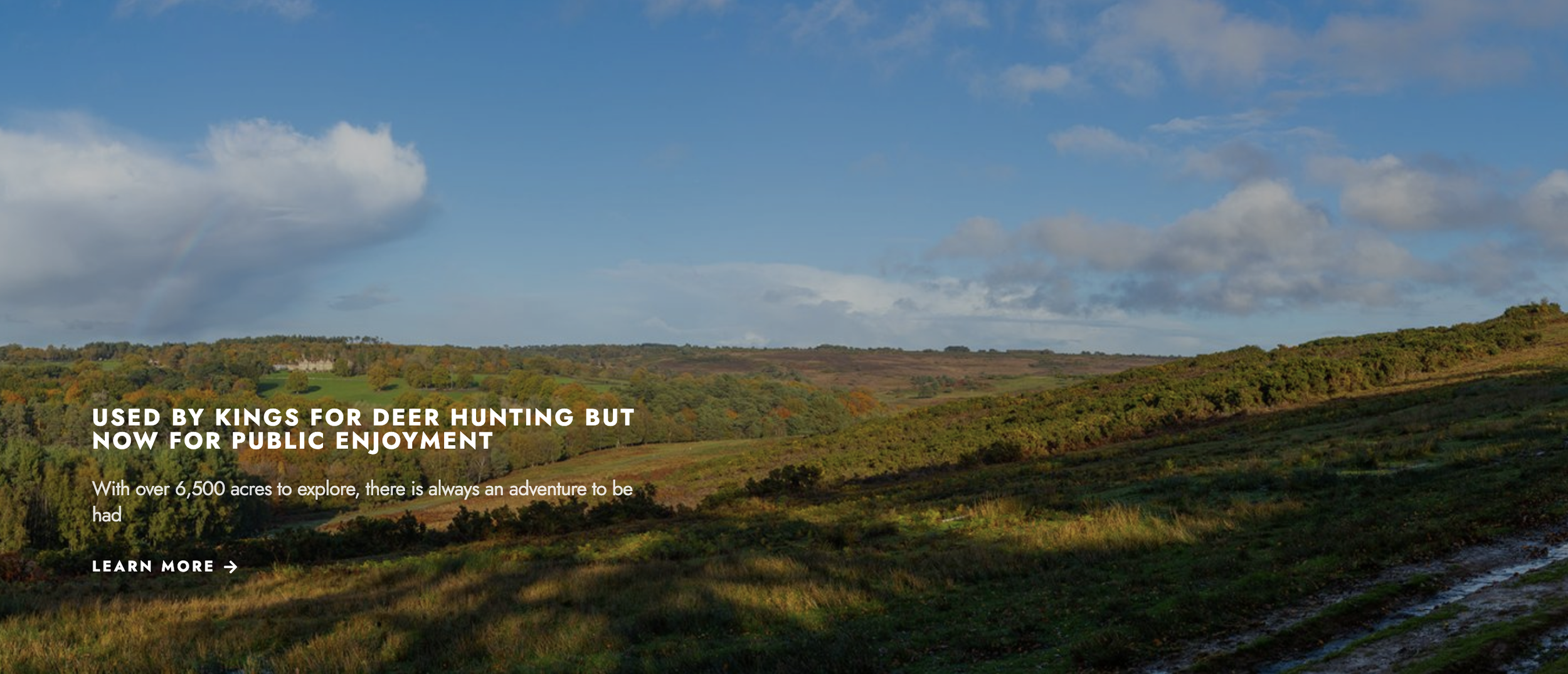 A scenic countryside landscape with rolling hills, green trees, and a partly cloudy sky with a rainbow in the distance.