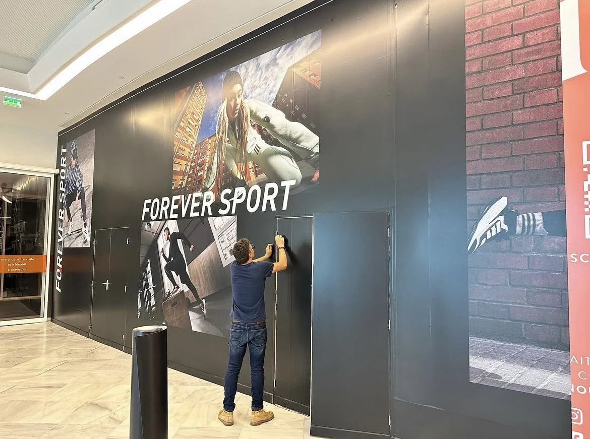 Un homme en train d'installer une publicité sur un mur noir dans un centre commercial. La publicité affiche des images de personnes en train de faire du sport et le texte « FOREVER SPORT ».